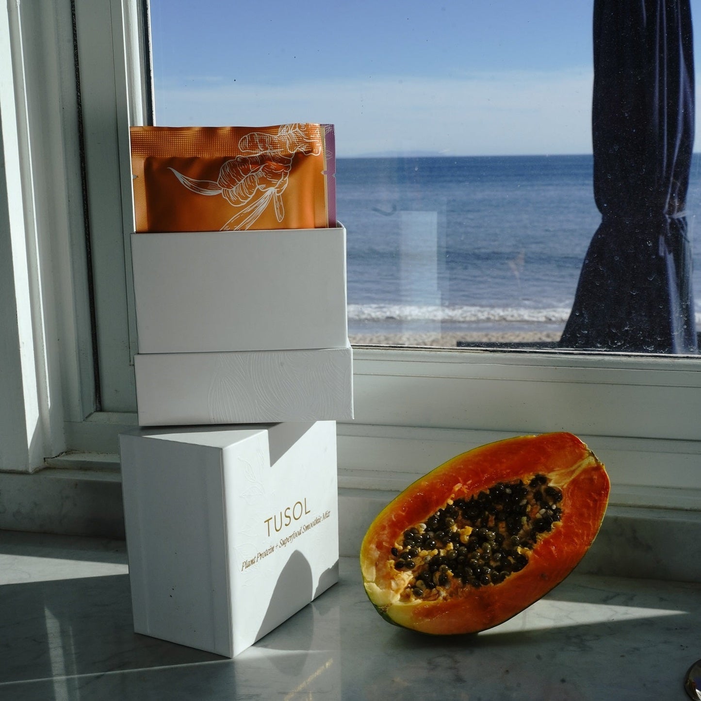 Plant Protein + Superfood Smoothie Mix sample box with organic smoothie packets next to halved papaya by the window overlooking the sea