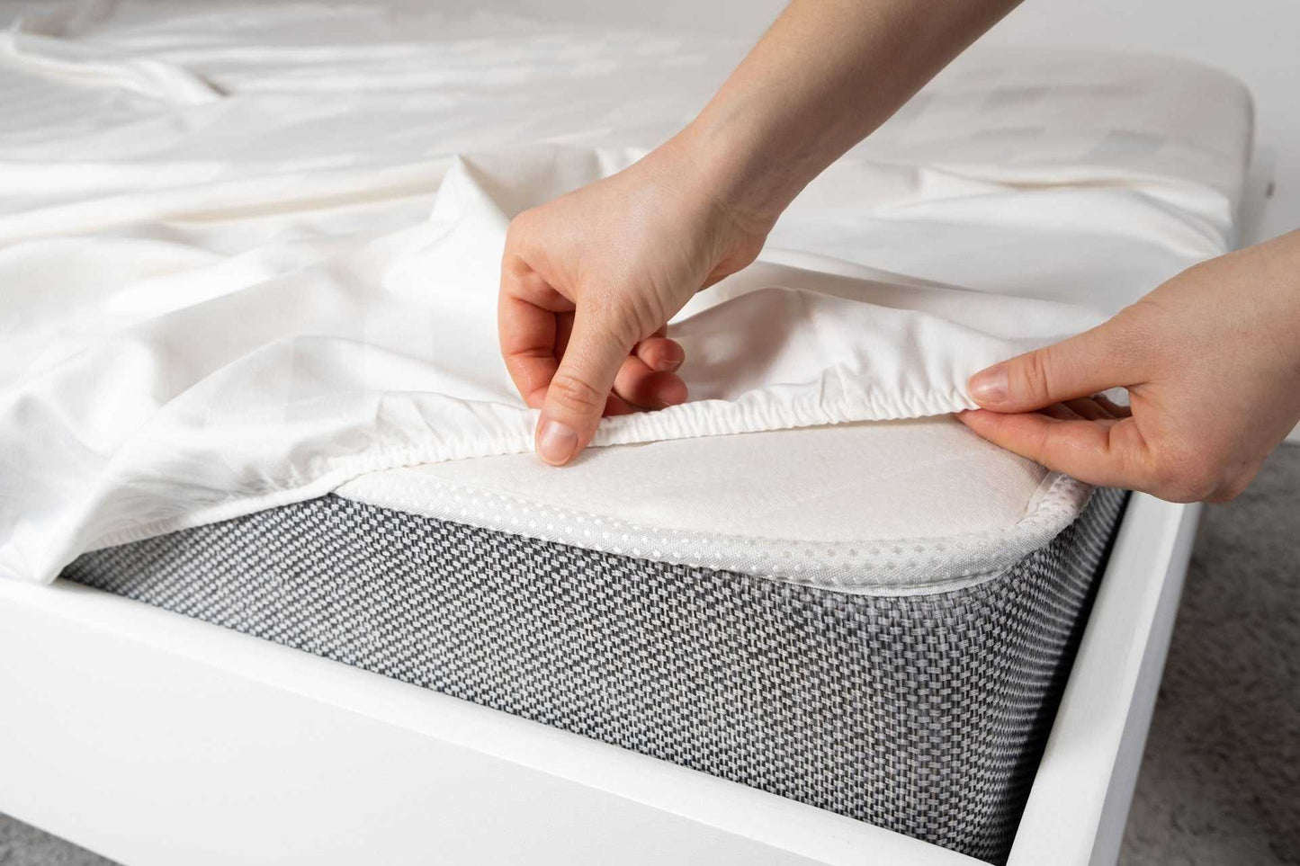 Hands adjusting white bedsheet over mattress with gray herringbone weave cotton blanket underneath