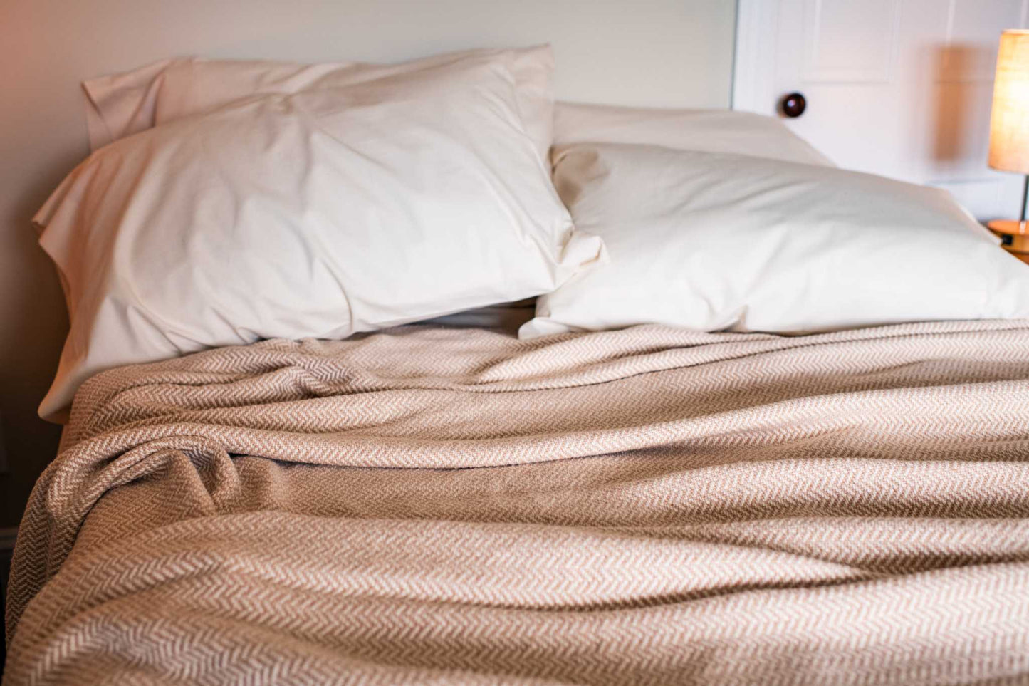 Herringbone weave cotton blanket in beige draped on bed with white pillows and warm lighting