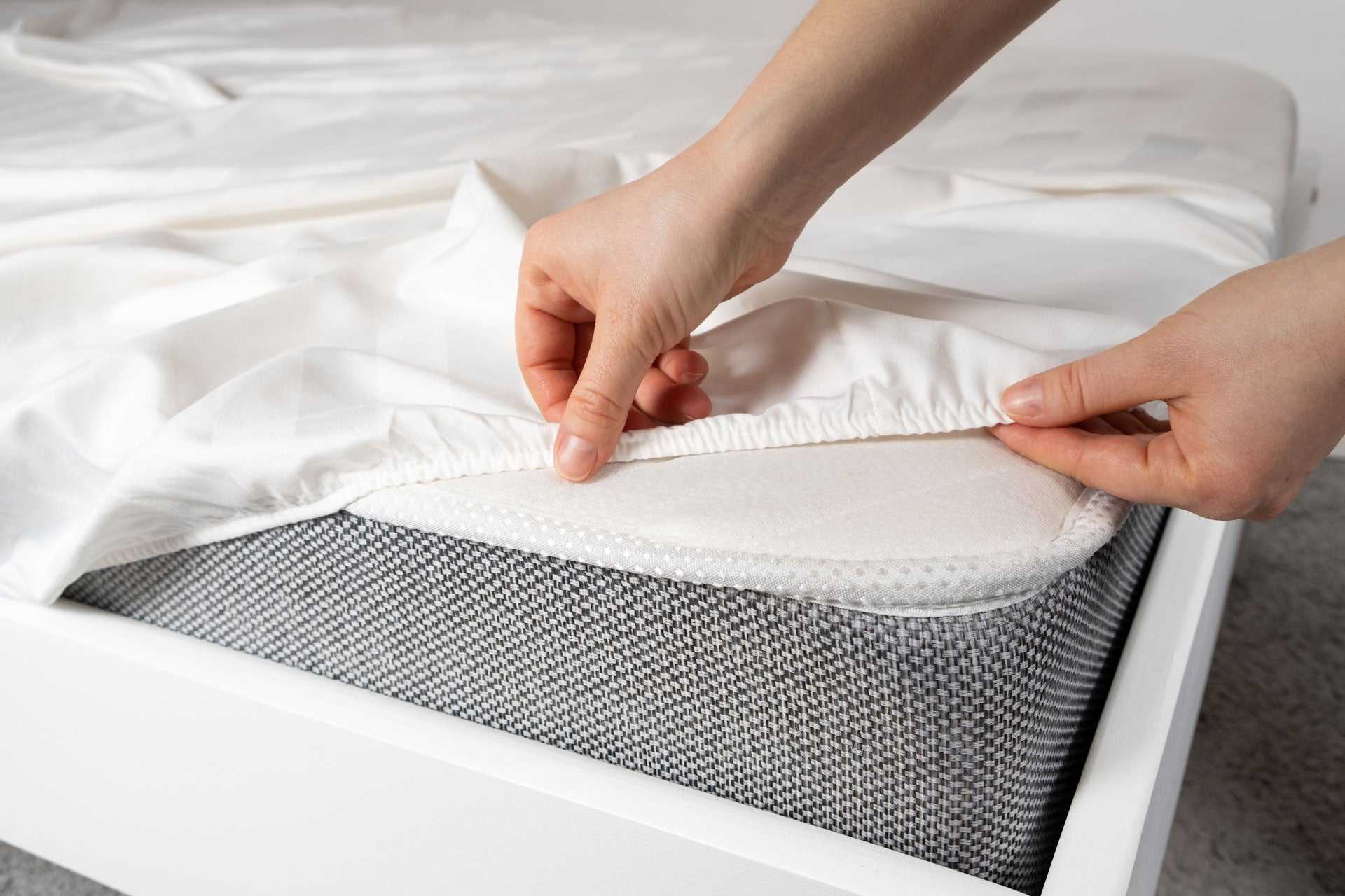 Hands adjusting white bedsheet over mattress with gray herringbone weave cotton blanket underneath