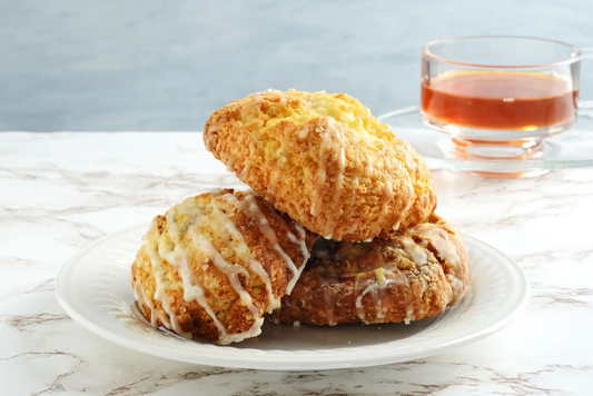 Three glazed scones including buttermilk scone on white plate with cup of tea in background