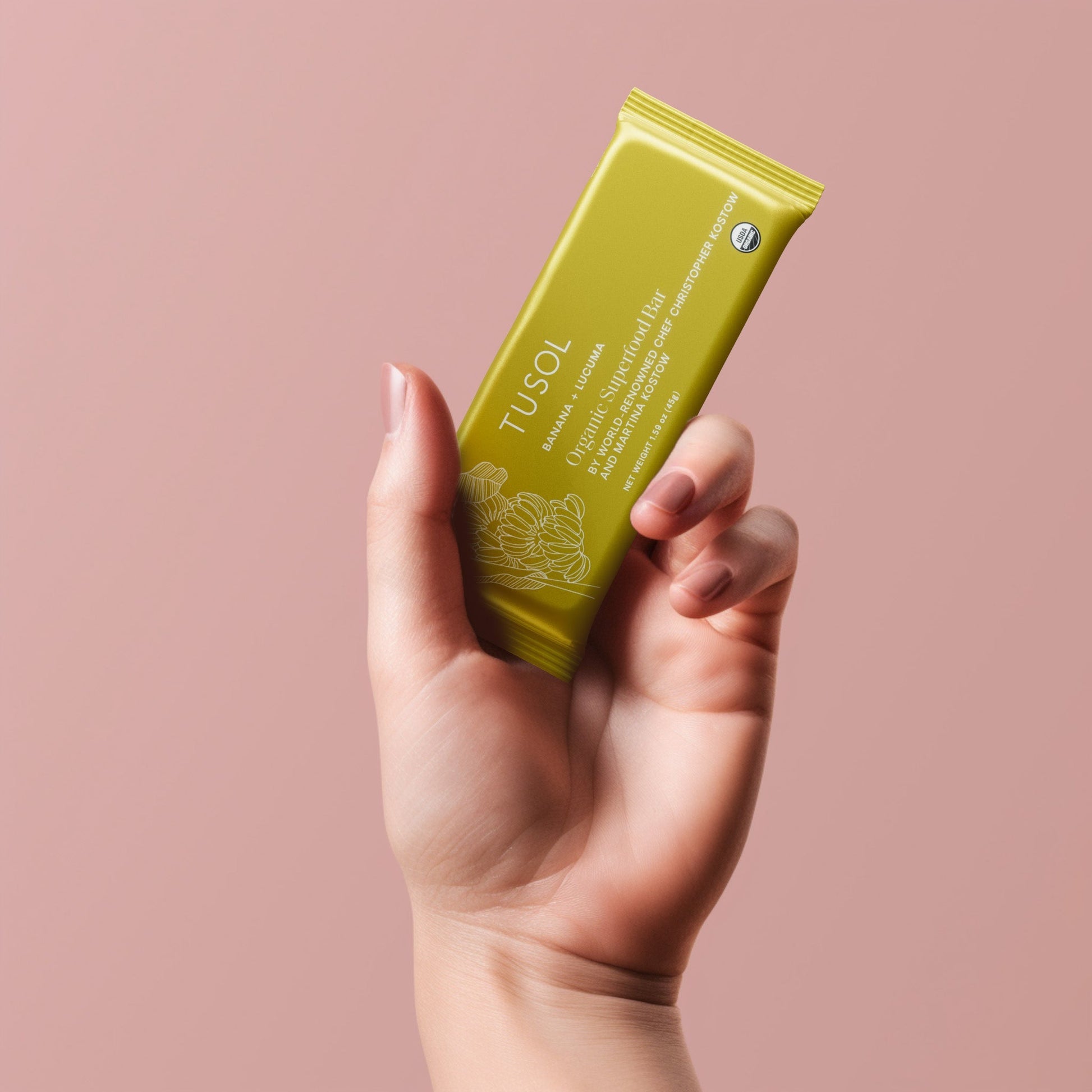 Hand holding a TUSOL Wellness Essentials Starter Kit organic protein bar with a pink background