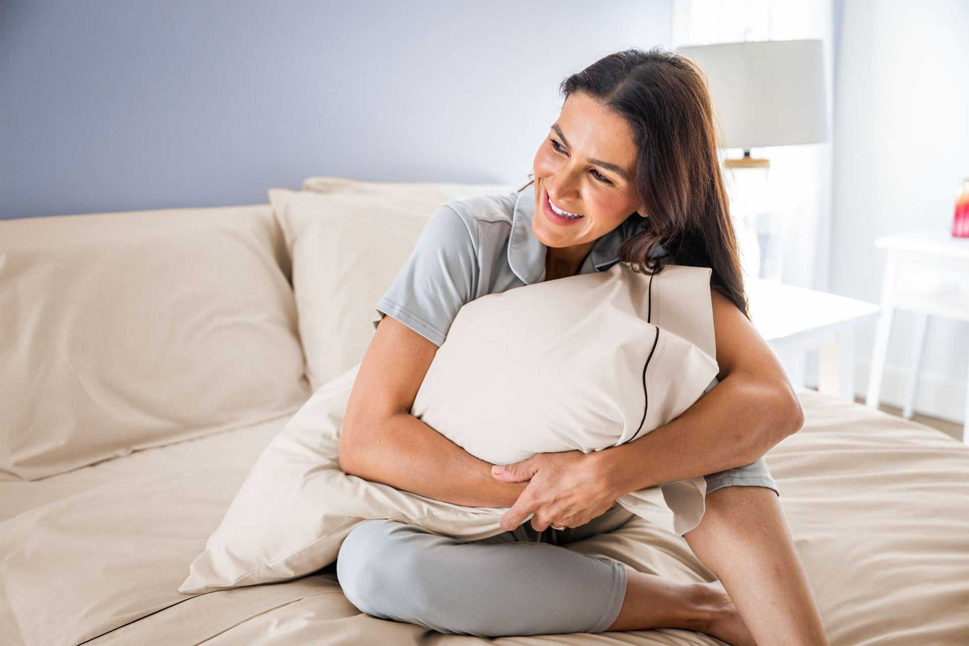Smiling woman hugging beige cotton piping design pillow on matching bed sheets in cozy bedroom