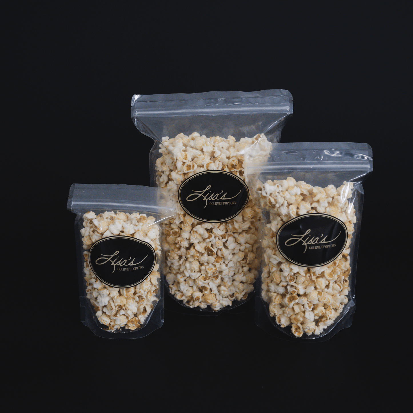 Three clear resealable bags of Kettlecorn popcorn in small, medium, and large sizes with black and gold Lisa's Gourmet Popcorn labels against a black background